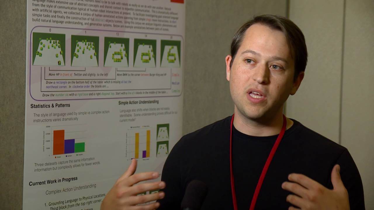 A photo of Yonatan Bisk standing in front of and explaining a poster behind him
