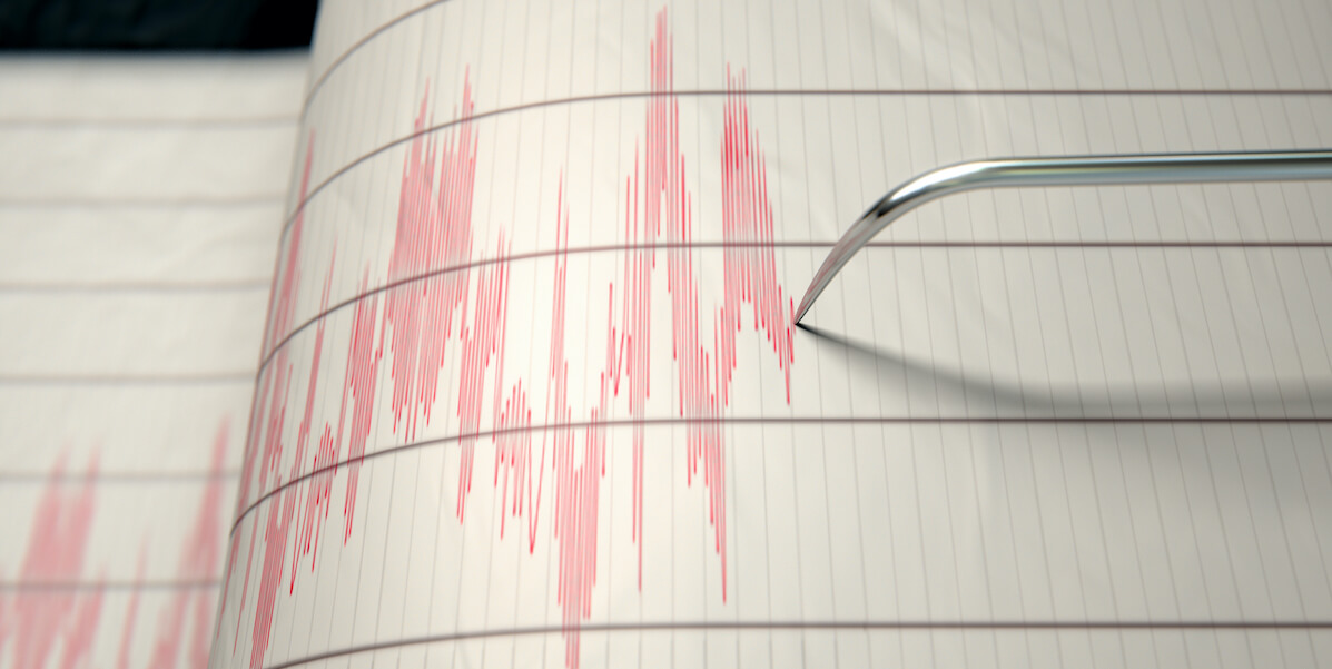 A closeup of a seismograph machine needle drawing a red line on graph paper depicting seismic and earthquake activity - Credit: Istock