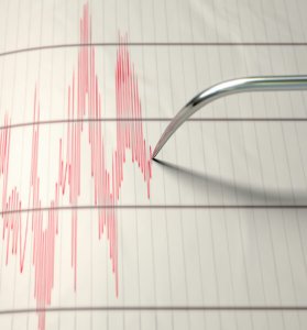 A closeup of a seismograph machine needle drawing a red line on graph paper depicting seismic and earthquake activity - Credit: Istock