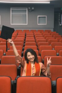 FU SITS IN A CLASSROOM AND WAVES HER GRADUATION CAP