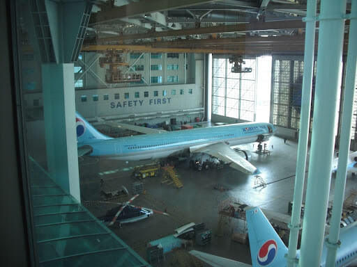 An indoor view of an aircraft maintenance facility with a large airplane bearing the Korean Air logo. The facility has high ceilings with overhead cranes and a 'Safety First' sign on the wall.