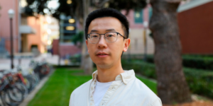 A man with glasses in a dress shirt standing in front of a garden.