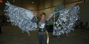 Image of Elisa McCormick dressed in a costume with a decorated silver top, thick black belt, and dark gray pants. A golden sash hangs from the belt. Elisa holds a pair of wings above her shoulders. Each wing is made out of long, acrylic and metal tubes with silver, reflective materials cut to look like feathers. On her head, Elisa wears a bronze hat placed sideways on her head. She is standing inside a warehouse with moderate lighting.