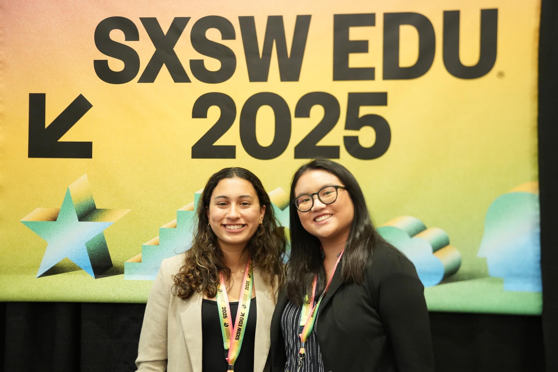Two girls smiling at the camera in front of a poster.