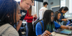 In the forefront of the image, a three people can be seen gathered in front of a desk. One person can be seen in the very left, looking to the right. One person stands in the center, looking towards an open laptop on the desk. Another person is maneuvering a mouse while looking at the open laptop in front of them. There is another person sitting next to the person with the laptop and they appear to be looking at their own laptop. The background setting is a classroom with numerous desks, a smart screen partially seen in the right corner, and a lit up window to the very left. In the distance, one person can be seen looking at the open laptop from further away with a sheet of paper in their hands. Two people can be seen in the very back of the room, one presumably looking at the smart board and the other is bending over to look at something on a desk.