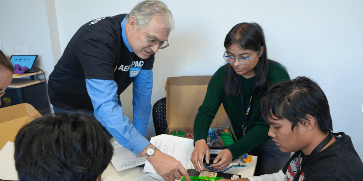 Four people can be seen gathered around a desk that is covered with papers, wires, and a circuit-making kit. One person is assembling the circuit in the kit (a green board with various rows of holes strung by red and black wires) with the support of two other people who appear to be gesturing at and rearranging pieces on the kit. Yellow, green, and black pieces are shown placed across the table. The fourth person is shown sitting at the table with a view of the back of their head, sitting on the opposite side of the three people working on the circuit kit. In the distance, to the left, a portion of a table with a small screen can be seen as well as the side of someone's head leaning towards the table.