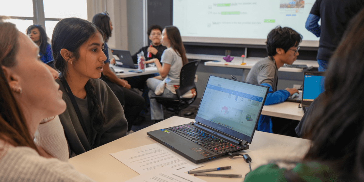 In the very front of a classroom setting (with a projector screen displaying instructions shown partially on the left and lit up windows with curtains to the right), we see three people sitting in front of an open laptop that appears to display coding software. The desk the laptop is placed on top of also has two sheets of paper and two pens on top. To the slight right, another table with one participant can be seen using a laptop. To the left, another desk can be seen with four participants can be partially pictured sitting around a laptop.