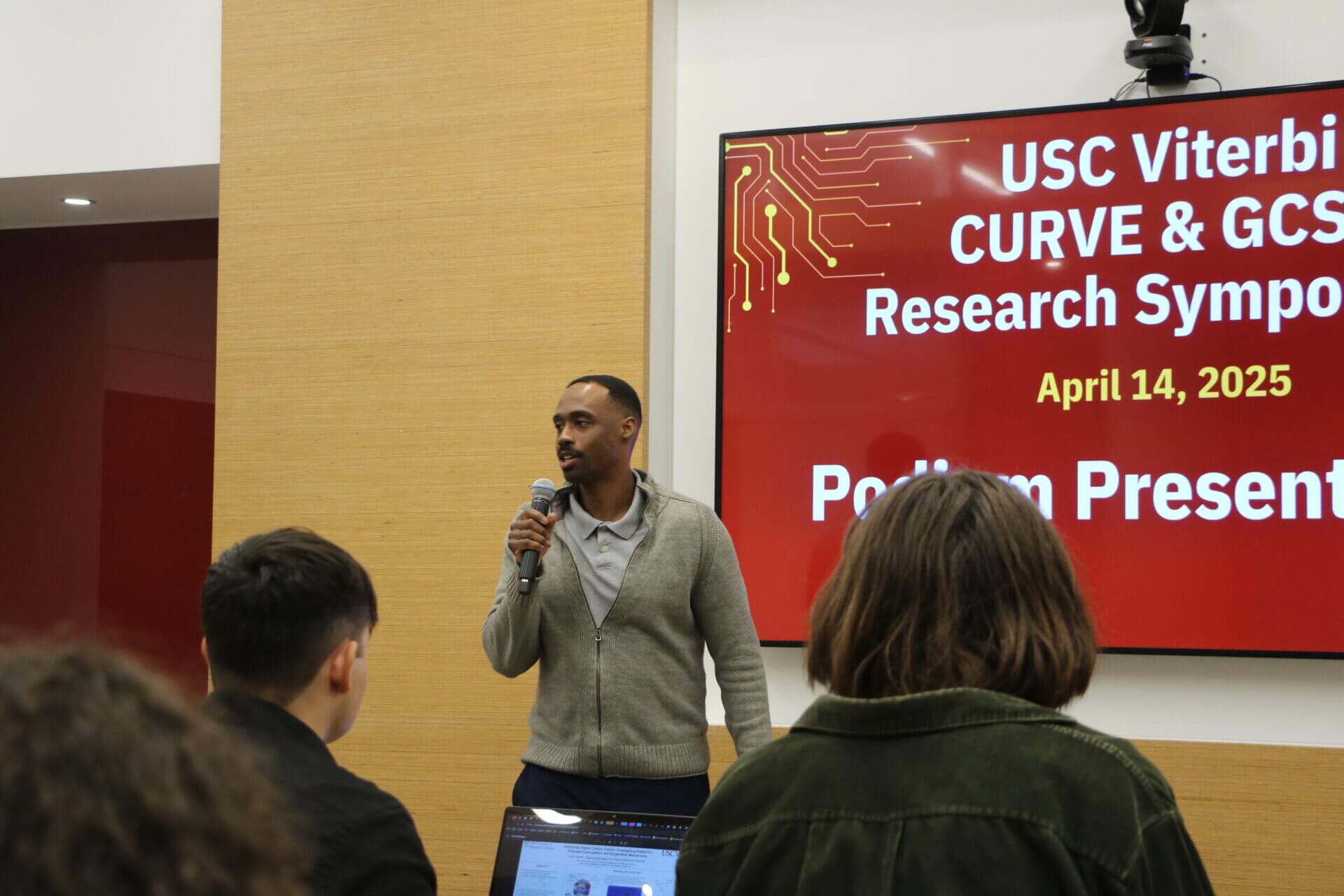 A man with a microphone addresses audience in front of a presentation title projected on a screen behind him