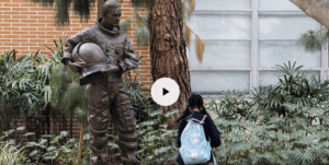 A play button for a video over an image of a girl looking at the statue of Neil Armstrong on USC's campus